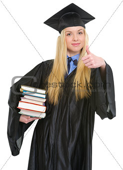 Happy young woman in graduation gown showing books and thumbs up
