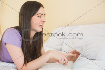 Cheerful brunette lying on a bed using a tablet pc in a bedroom