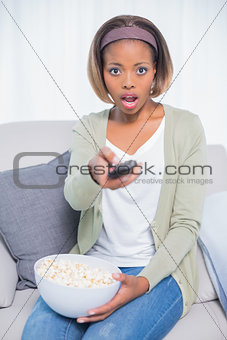 Shocked woman sitting on sofa changing tv channel while holding popcorn