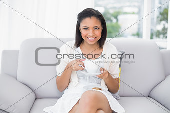 Pleased young dark haired woman in white clothes enjoying coffee and cookies