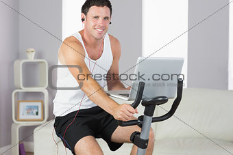 Sporty smiling man with earphones exercising on bike and holding laptop