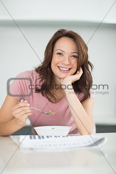 Smiling woman with bowl of cereals reading newspaper in kitchen