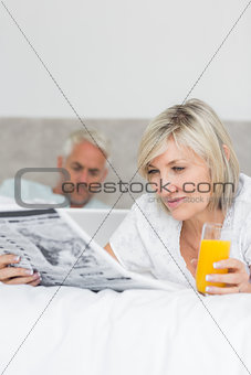 Couple reading newspaper and using laptop in bed