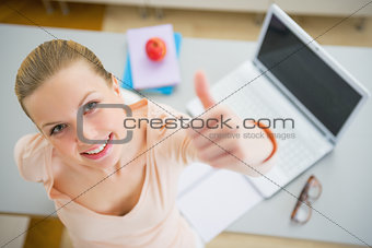 Happy young woman with books and laptop in kitchen showing thumb