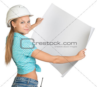 Beautiful woman builder looking at camera and holding in front of him large paper sheet. Rear view