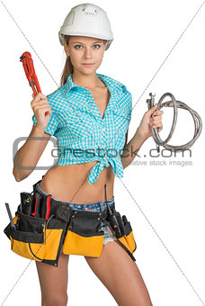 Pretty girl in helmet, shorts, shirt, tool belt with tools holding flexible hose and wrench