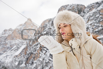 Woman in white coat and fur hat blowing snow from hand outdoors