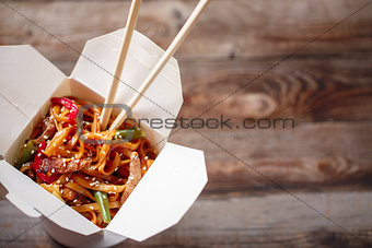 Noodles with pork and vegetables in take-out box on wooden table