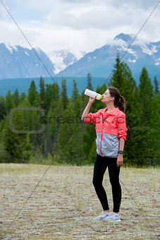 Beautiful woman runner drinking water outdoors. Mountains background.