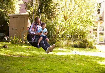 Father And Son Having Fun On Tire Swing In Garden