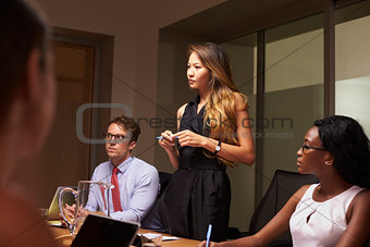Businesswoman stands among seated team at an evening meeting