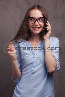 portrait of smiling woman with glasses speaking by cellphone near grey background wall