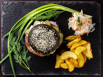 Black hamburger with onions and cucumbers on stone table with black background.