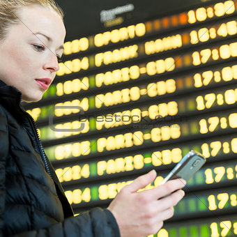 Woman at airport in front of flight information board checking her phone.