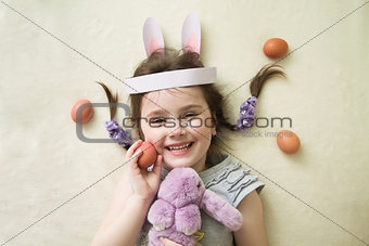 a little smiling girl in a bunny's paper ears posing with a lilac bunny toy and a brown egg as symbols of Easter