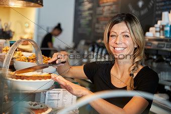 Waitress Behind Counter In Coffee Shop Cutting Slice Of Cake