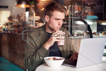 Young Man Using Laptop In Cafe Whilst Eating Breakfast