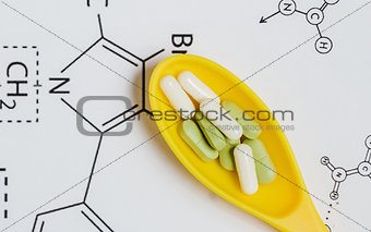 Assortment of Pills, Tablets and Capsules in Yellow Spoon on White Background with Chemical Formula.