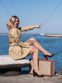 Attractive Young Blonde Woman in Trench with Vintage Suitcase is Sitting on the Jacht Pier and point with a finger in the Sea