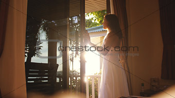 Pretty girl opens the door in the early morning during sunrise with lens flare effects and go out to the terrace